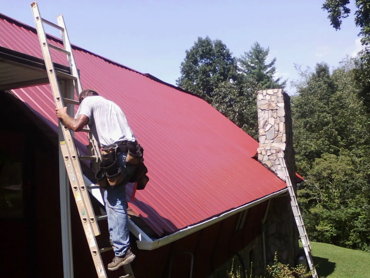 Roofer on a ladder beside a red metal roof and stone chimney — Soffit & Fascia Repair by Roofing Brothers in Addy